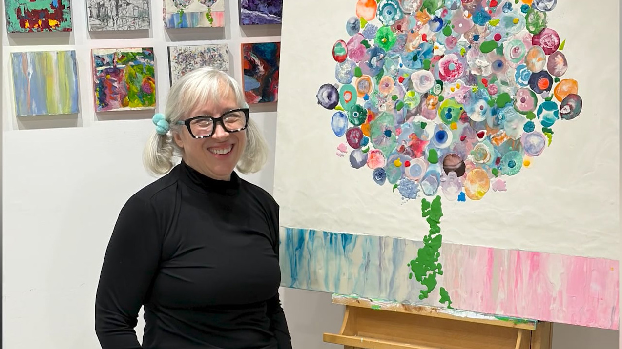 This image shows a photo of Karen Holloway smiling standing next to her Tree of Tears painting.